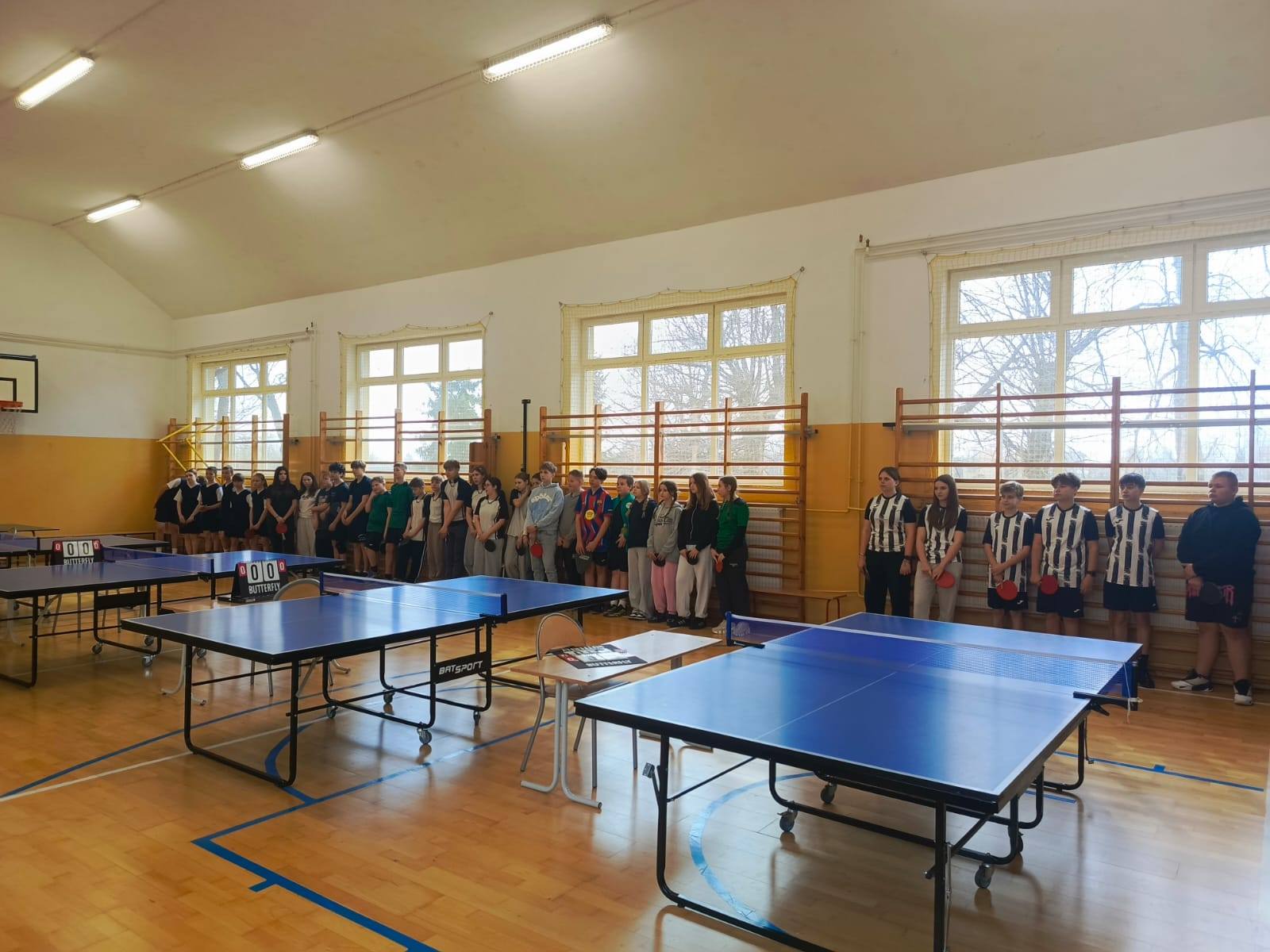 Uczniowie ustawieni w sali gimnastycznej przy stołach do tenisa stołowego podczas rozpoczęcia turnieju.